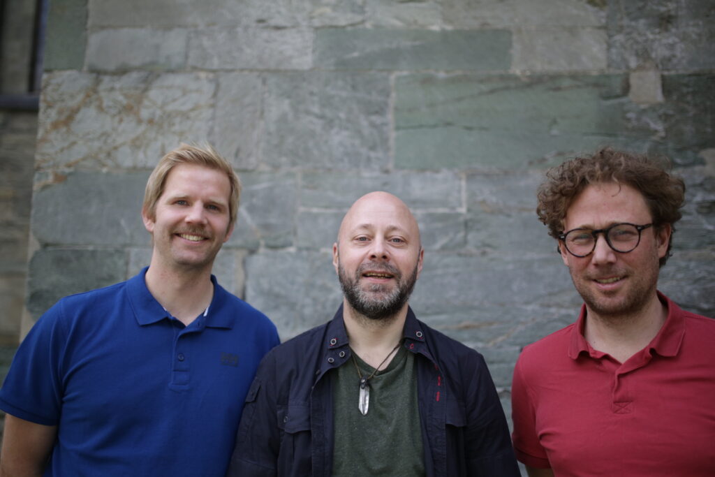 Portrettbilde av Håvard Wettland Gossé, Gjermund Gisvold og regissør Leiv Igor Devold foran en murvegg