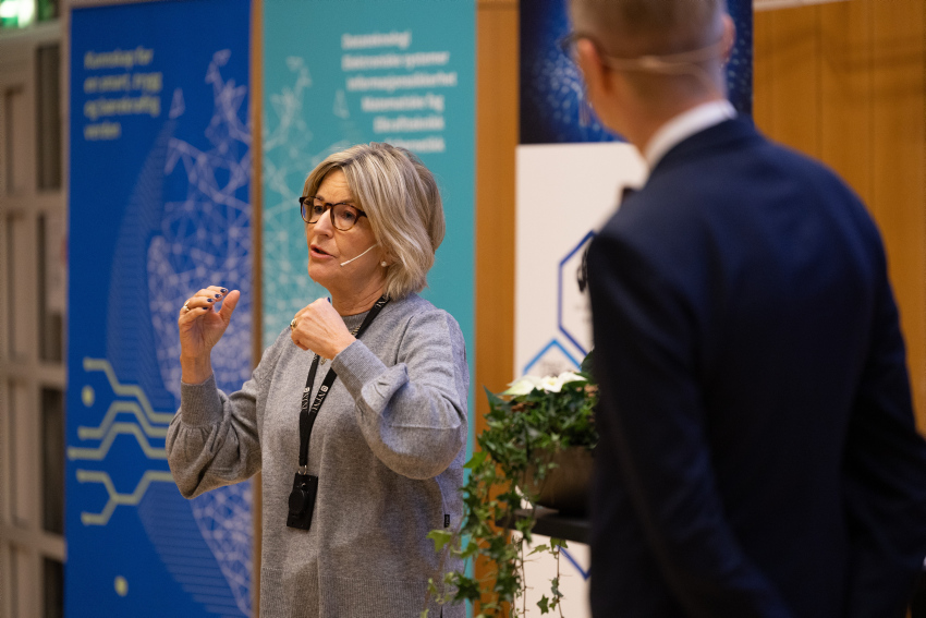 Ingrid Schjølberg står på en scene og snakker og gestikulerer mot publikum. Det står en mann på scenen og lytter til henne. Bak henne er store plakater for Fakultet for informasjonsteknologi og elektronikk og norwegian open AI lab.