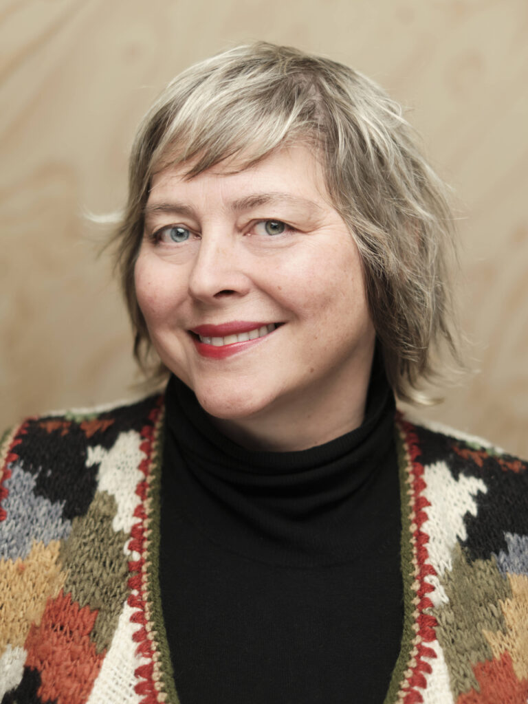 Bilde av Mari Nilsen Neira. Hun smiler til kamera, og har på seg en fargerik cardigan, Hun har kort, blondt hår.