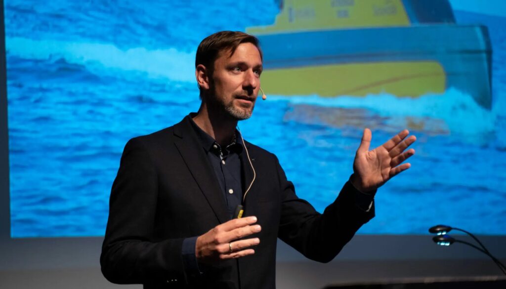 Bilde av professor Ole Andreas Alsos. Han har på seg en mygg, og står på en scene. Bak han er det et prosjektorbilde av vann og en liten skute. Han har på seg en sort dressjakke.